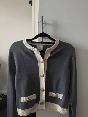 Grå cardigan med vita detaljer - Stilren grå cardigan med vita ribbade detaljer vid krage, knappslå, nederkant. Guldknappar framtill och dekorativa fickor ger en klassisk look. Perfekt att matcha med jeans eller kjol för en snygg outfit. Storlek xs