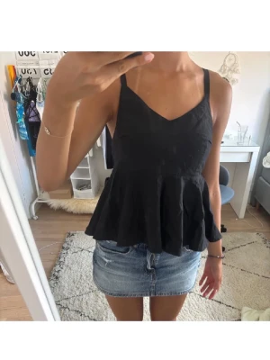 Svart volangtop med knyt i ryggen - Snygg svart linnetopp med volang nertill och smala axelband. Toppen har v-ringning framtill och knytning i ryggen för en extra detalj. Perfekt till jeans eller kjol för en trendig look.