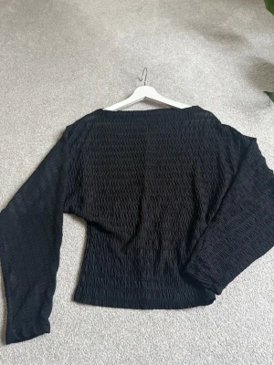 Svart långärmad topp med struktur - Säljer en svart långärmad topp med snygg strukturerad yta och lös passform. Toppen har bred halsringning och är tillverkad i ett lätt, stretchigt material som ger en cool och modern look. Perfekt att styla med jeans eller kjol.