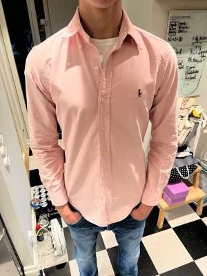 Rosa skjorta från Ralph Lauren - Säljer en klassisk rosa skjorta från Ralph Lauren med button-down krage och broderad logga på bröstet. Skjortan har långa ärmar, vita knappar och är tillverkad i ett mjukt bomullstyg. Perfekt för en stilren och avslappnad look.