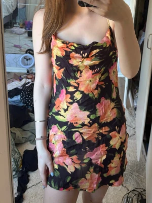 Blommig kort klänning med smala band - Snygg kort klänning med svart botten och stora blommor i orange, rosa och gul. Klänningen har smala axelband och en lätt figurnära passform. Perfekt för varma dagar och sommarens alla tillfällen. Materialet känns lätt och luftigt! 💓🌸