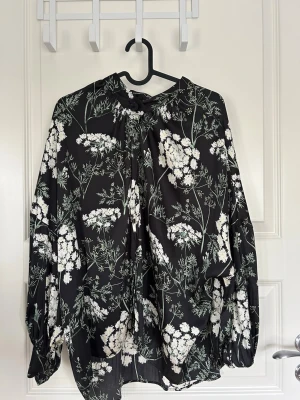 Svart blommig blus med knytning - Svart blus med vita och gröna blommor, lös passform och knytning vid halsen. Långa ärmar med lätt puff och knappdetalj vid halsringningen. .