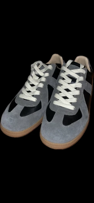 Maison Margiela svarta och grå sneakers - Snygga sneakers från Maison Margiela i svart läder med grå mockadetaljer och beige/brun sula. Klassisk låg modell med vita snören och stilren design som passar till det mesta. Perfekta för dig som gillar exklusiva och trendiga skor.