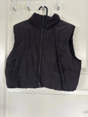 Svart dunväst med hög krage -  Svart dunväst med dragkedja framtill och två fickor på sidorna. Västen har en hög krage och en puffig, quiltad design.