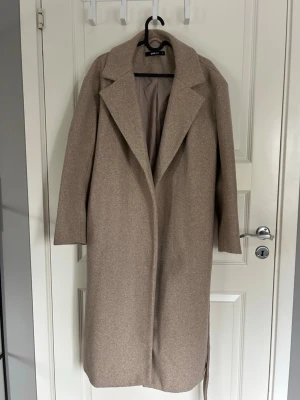Beige kappa med bälte från Gina Tricot -  beige kappa från Gina Tricot.  Kappan har lång ärm och rak passform, perfekt för höst och vinter. I storlek S