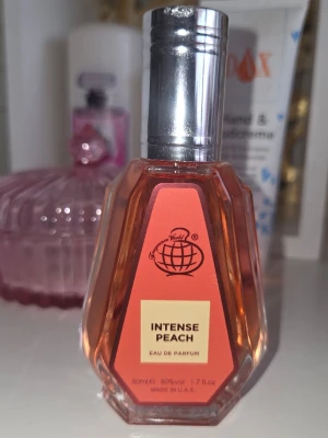 Intense Peach Eau de Parfum - En snygg parfymflaska från Fragrance World med doften Intense Peach. Flaskan är facetterad och har en persikofärgad etikett samt silverfärgad kork. Innehåller 50 ml eau de Parfum 