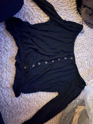 Svart långärmad topp Stories for her - Svart långärmad topp från Stories for her med dekorativa knappar längs framsidan. Tunn och mjuk i materialet, perfekt till jeans eller kjol. Enkel och stilren design som funkar till många olika looks.