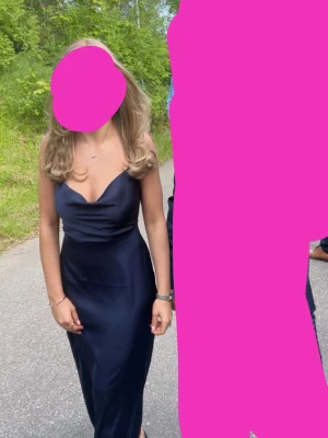 Marinblå långklänning i satin - Balklänning! Endast använd en gång på min bal. Marinblå långklänning med smala axelband och djup v-ringning. Klänningen har en figurnära passform och slät, glansig yta som ger en lyxig känsla. Perfekt för speciella tillfällen.