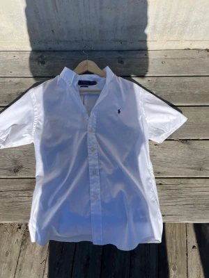 Vit kortärmad skjorta Polo Ralph Lauren - Klassisk vit kortärmad skjorta från Polo Ralph Lauren i slim fit. Skjortan har button-down krage, knappar framtill och den ikoniska broderade loggan på bröstet. Tillverkad i bomull för en fräsch och stilren look.