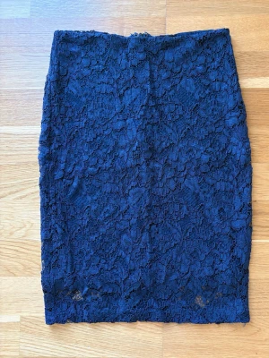 Mörkblå spetskjol i figurnära modell från Stockh LM - Snygg mörkblå kjol med figurnära passform och vacker spets över hela plagget. Kjolen är kort och har en elegant, feminin look som passar perfekt till olika stilar. Ingen synlig dragkedja eller knappar, bara ren spetsdesign. Längd 53 cm.