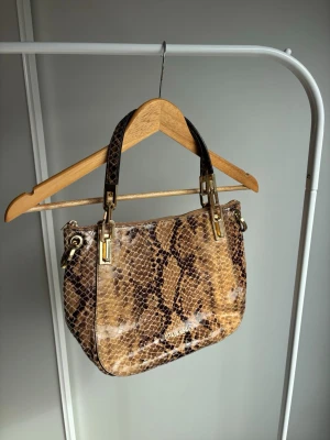 Michael Kors ormmönstrad handväska - Snygg handväska från Michael Kors med brunt och beige ormmönster. Väskan har guldfärgade metalldetaljer och två rejäla axelband. Tillverkad i glansigtkonstläder med präglad struktur för en lyxig känsla.
