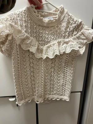 Beige spetsblus med volang från Zara - Supersöt beige spetsblus från Zara med korta ärmar och broderad volangdetalj över bröstet och axlarna. Blusen har hög krage med volangkant och ett vackert hålmönster i hela tyget. Perfekt för dig som gillar romantisk och vintageinspirerad stil.