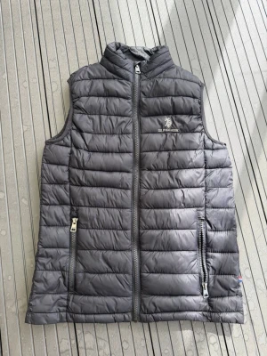  U.S. Polo Assn. - Snygg svart dunväst från U.S. Polo Assn. med quiltad design och dragkedja framtill. Västen har två praktiska fickor med dragkedja och en hög krage. Perfekt för lager-på-lager och stilren look. Hör av dig vid frågor och funderingar ☺️