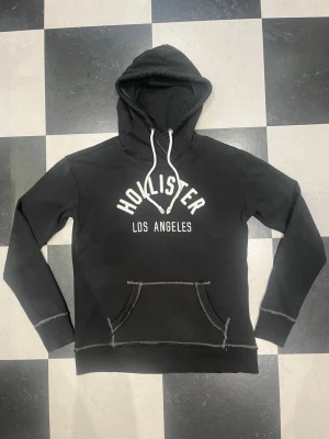 Svart Hollister hoodie med tryck - Svart hoodie från Hollister med vit text 'Hollister Los Angeles' på bröstet. Klassisk känguruficka framtill, vita dragsnören i huvan och synliga sömmar i kontrastfärg. Perfekt för en avslappnad streetstyle-look.