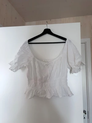 Vit blus med volang och puffärm - Söt vit blus med volangkant och puffiga korta ärmar. Modellen har smock i midjan och en liten knytning framtill. Perfekt för en romantisk look och passar till både jeans och kjol.