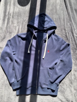 Zip Hoodie Polo Ralph Lauren - Snygg marinblå hoodie från Polo Ralph Lauren med dragkedja, vit dragsko och röd broderad logga på bröstet. Mjuk insida och känguruficka framtill. Perfekt för en chill och stilren look.