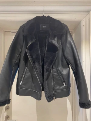 Svart skinnjacka med fuskpäls - Snygg svart skinnjacka från DeFacto med mjukt fuskpälsfoder. Jackan har dragkedjor på fickorna och asymmetrisk dragkedja framtill. Perfekt för dig som vill ha en cool och trendig look med extra värme.