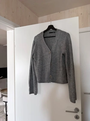 Grå kofta med knappar - Mysig grå kofta med v-ringning och tre knappar framtill. Koftan har långa ärmar och är stickad i ett mjukt material. Perfekt att slänga på sig när det är lite kyligt ute.