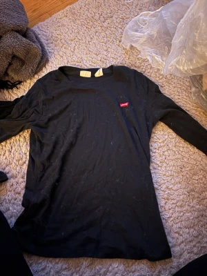 Svart långärmad topp från Levi's - Svart långärmad topp från Levi's med klassisk rund halsringning och den ikoniska röda Levi's-loggan på bröstet. Tillverkad i mjukt bomullsmaterial som är skönt mot huden. Perfekt för en avslappnad och stilren look.