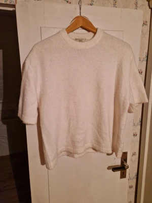 Vit stickad t-shirt från Gina Tricot - Säljer en vit stickad t-shirt från Gina Tricot i premiumkvalitet. Tröjan har korta ärmar, rund halsringning och ribbstickade kanter. Perfekt för dig som gillar en enkel och stilren look med en twist av stickat.