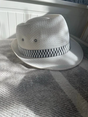 Vit fedora med rutigt band - Snygg vit fedora med svartvitt rutigt band runt kronan. Hatten har två metallöljetter på sidan för ventilation och är tillverkad i ett lätt, strukturerat material. Perfekt för att lyfta din stil med en cool accessoar.storlek 57-58