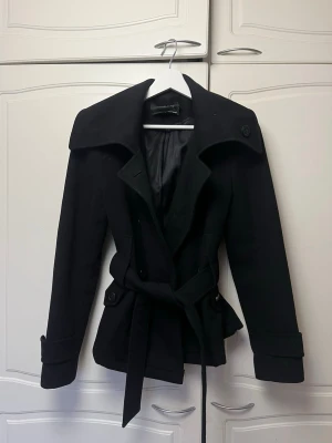 Svart trenchcoat med bälte - Stilren svart trenchcoat med bred krage och knytskärp i midjan. Jackan har knappar vid ärmslut och axlar samt slät insida. Perfekt för dig som vill ha en klassisk och tidlös look. Jackan är förkortad och det syns på insidan, men ej på utsidan eller vid annvändning  (se sista bilden)