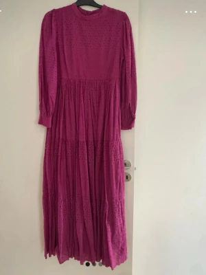 Lång lila klänning med broderade detaljer - Lång lila klänning med broderade hål och lång ärm. Klänningen har rund halsringning och ett luftigt, böljande fall. Perfekt för dig som gillar romantiska och färgstarka plagg. Den är äkta bomull