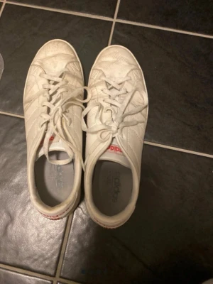 Vita Adidas sneakers med röda detaljer - Snygga vita sneakers från Adidas med klassiska röda detaljer och logga på hälen. Skorna har lågt skaft, snörning och är tillverkade i läderliknande material. Perfekta för en stilren och sportig look.