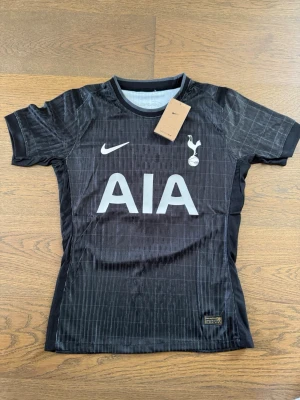 Svart Tottenham matchtröja Nike - Snygg svart Tottenham Hotspur matchtröja från Nike med AIA-logga på bröstet och klubbmärke. Tröjan har XAVI och nummer 7 på ryggen samt korta ärmar. Tillverkad i lätt och ventilerande material, perfekt för fotbollsträning eller match.