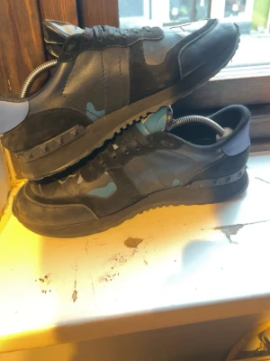 Valentino Garavani svarta sneakers - Snygga svarta sneakers från Valentino Garavani med blå och grå kamouflagedetaljer. Skorna har lågt skaft, svarta snören och coola nitar på sulan. Materialmix av läder och textil ger en exklusiv känsla. Perfekta för dig som vill sticka ut med stil.