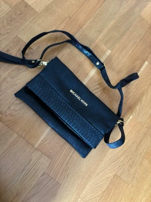 Svart axelväska Michael Kors - Snygg svart axelväska från Michael Kors i präglat konstläder med guldfärgade detaljer. Väskan har justerbar axelrem, magnetlås och flera fack, inklusive ett med dragkedja. Perfekt för att bära det viktigaste med stil. Väskan är inte äkta!