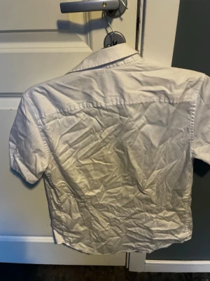Vit kortärmad skjorta från Guess - Snygg vit skjorta från Guess med korta ärmar och klassisk krage. Skjortan har knappar framtill och är tillverkad i bomull, vilket gör den bekväm och lätt att bära. Perfekt för en stilren och fräsch look.