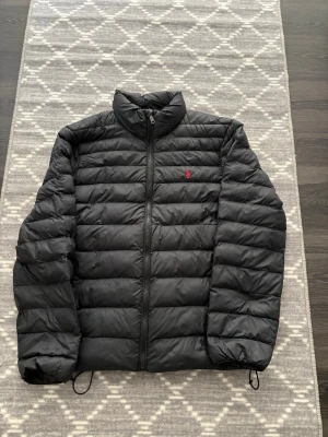 Svart pufferjacka från Polo Ralph Lauren - Svart pufferjacka från Polo Ralph Lauren med röd broderad logga på bröstet. Jackan har dragkedja framtill, ståkrage och justerbar dragsko nedtill. Perfekt för kalla dagar och riktigt skön att bära. Klassisk quiltad design och lätt vaddering. (Litet märke vid bilden som knappas syns)