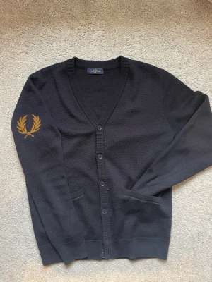 Fet Fred Perry cardigan - Mörkblå Fred Perry kofta med logga på ena axeln. Toppskick/ som ny, älskar tröjan. Gjord av 50% ull och 50% akryl som ger en merinoull effekt och inte kliar. Är i storlek M och anser att den är normal i storleken. Ett plagg som skriker hög kvalitet och fin detalj.