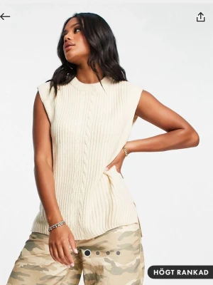 Ärmlös stickad väst i offwhite - Snygg ärmlös stickad väst i offwhite med kabelstickat mönster framtill. Rund halsringning och avslappnad passform. Köpt på Asos, passar S 