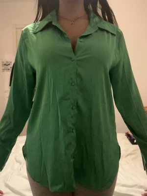 Grön silky skjorta med knappar - Snygg grön skjorta med klassisk krage och knappar framtill. Skjortan har lång ärm och är tillverkad i ett mjukt, lätt material som ger en avslappnad passform. Perfekt att styla både uppknäppt och stängd.