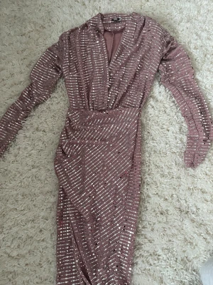 Glittrig rosa långklänning med paljetter - Snygg långklänning i rosa med glittriga paljetter över hela klänningen. Djup v-ringning och långa ärmar ger en elegant look. Perfekt för dig som vill sticka ut med en glamorös stil.