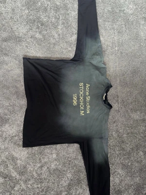 Svart långärmad topp Acne Studios - Cool långärmad topp från Acne Studios med Stockholm 1996-tryck på bröstet. T-shirten har en svart färg med en gradient-effekt mot grått och rund halsringning. Perfekt för dig som gillar stilrena och trendiga plagg.
