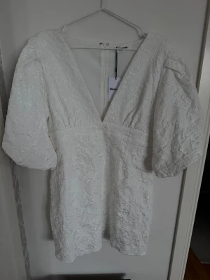 Vit kort klänning från Showpo - Supersnygg vit kort klänning från Showpo med djup v-ringning och puffiga korta ärmar. Klänningen har ett broderat mönster i tyget och markerad midja. Dragkedja bak för enkel påklädning. Säljare då jag köpte fel storlek.