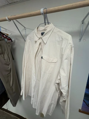 Randig skjorta från Ralph Lauren - Säljer en klassisk långärmad skjorta från Ralph Lauren i ljusbeige och vit med tunna ränder. Skjortan är i 100% bomull och har bröstficka med logotyp, knappar framtill och en snygg krage. Perfekt för dig som gillar stilrena plagg. Storlek XXL