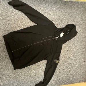 Svart hoodie från Stone Island - Säljer en svart hoodie från Stone Island med dragkedja och huva. Klassisk logopatch på vänster ärm och praktiska fickor framtill. Materialet är mjukt och bekvämt, perfekt för en avslappnad streetwear-look.
