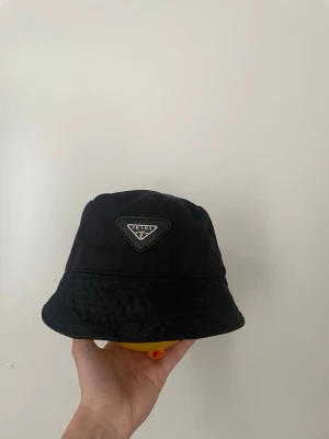 Svart bucket hat från Prada - Snygg svart bucket hat från Prada med det klassiska trekantiga logomärket framtill. Hatten är tillverkad i ett glansigt nylonmaterial som ger en modern och clean look. Perfekt accessoar för att lyfta din outfit.