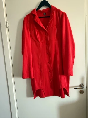 Röd långärmad skjortklänning - Röd skjortklänning med långa ärmar och markerad byst, knappar framtill och krage. Klänningen har en kort längd och lätt figurnära passform. Perfekt för dig som vill sticka ut med en färgstark look.