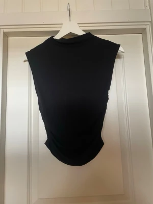 Svart ärmlös topp med hög hals - Säljer en svart, ärmlös topp med hög hals och tight passform. Perfekt att styla med jeans eller kjol för en clean och modern look. Materialet känns mjukt och stretchigt, vilket gör den bekväm att bära hela dagen.