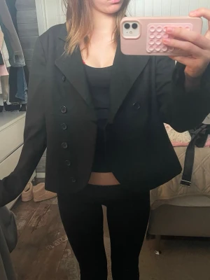 Svart kort kavaj med dubbelknäppning - Snygg svart kavaj med dubbelknäppning och breda slag. Modellen är kortare och har en boxig passform som ger en trendig look. Perfekt att styla över en topp eller t-shirt för en cool outfit. Materialet känns slätt och klassiskt. Passar xs-m beroende pä önskad passform!