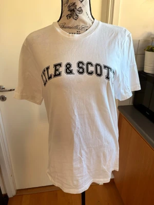 Vit Lyle & Scott t-shirt med tryck - Vit t-shirt från Lyle & Scott med svart texttryck över bröstet. Klassisk rund halsringning och korta ärmar. Perfekt basic-plagg med en clean och sportig vibe.