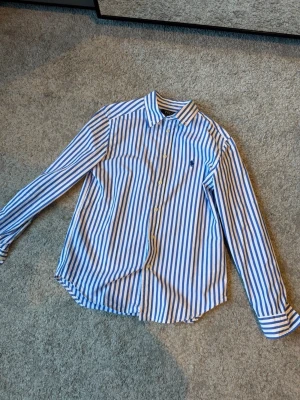 Blåvit randig skjorta från Ralph Lauren - Snygg blåvit randig skjorta från Ralph Lauren använt två gånger med klassisk krage och långa ärmar. Skjortan har knappar framtill och den ikoniska broderade loggan på bröstet. Perfekt för en stilren look.