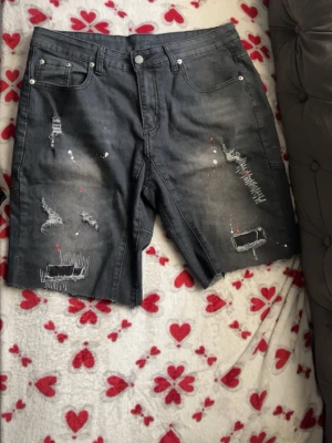 Grå slitna jeansshorts med detaljer - Säljer ett par grå jeansshorts med slitningar, trasiga detaljer och färgstänk i vitt och rött. Shortsen har fem fickor och en klassisk jeanslook med knapp och dragkedja. Perfekta för en avslappnad och trendig stil.