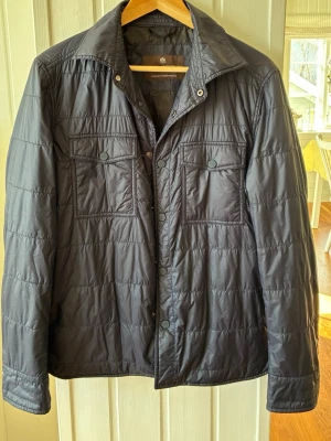 Mörkblå vindjacka från Oscar Jacobson - Snygg mörkblå vindjacka från Oscar Jacobson med två bröstfickor med knappstängning och klassisk krage. Jackan har tryckknappar framtill och är tillverkad i ett lätt, glansigt material som passar perfekt för blåsiga dagar.