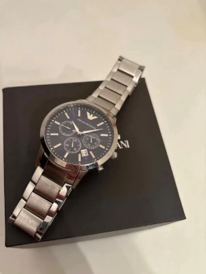 Emporio Armani silverklocka herr - Stilren herrklocka från Emporio Armani med silverfärgad länk i metall och mörkblå urtavla. Klockan har tre mindre urtavlor, datumvisning och klassisk logga upptill. Perfekt accessoar för en snygg och modern look. 